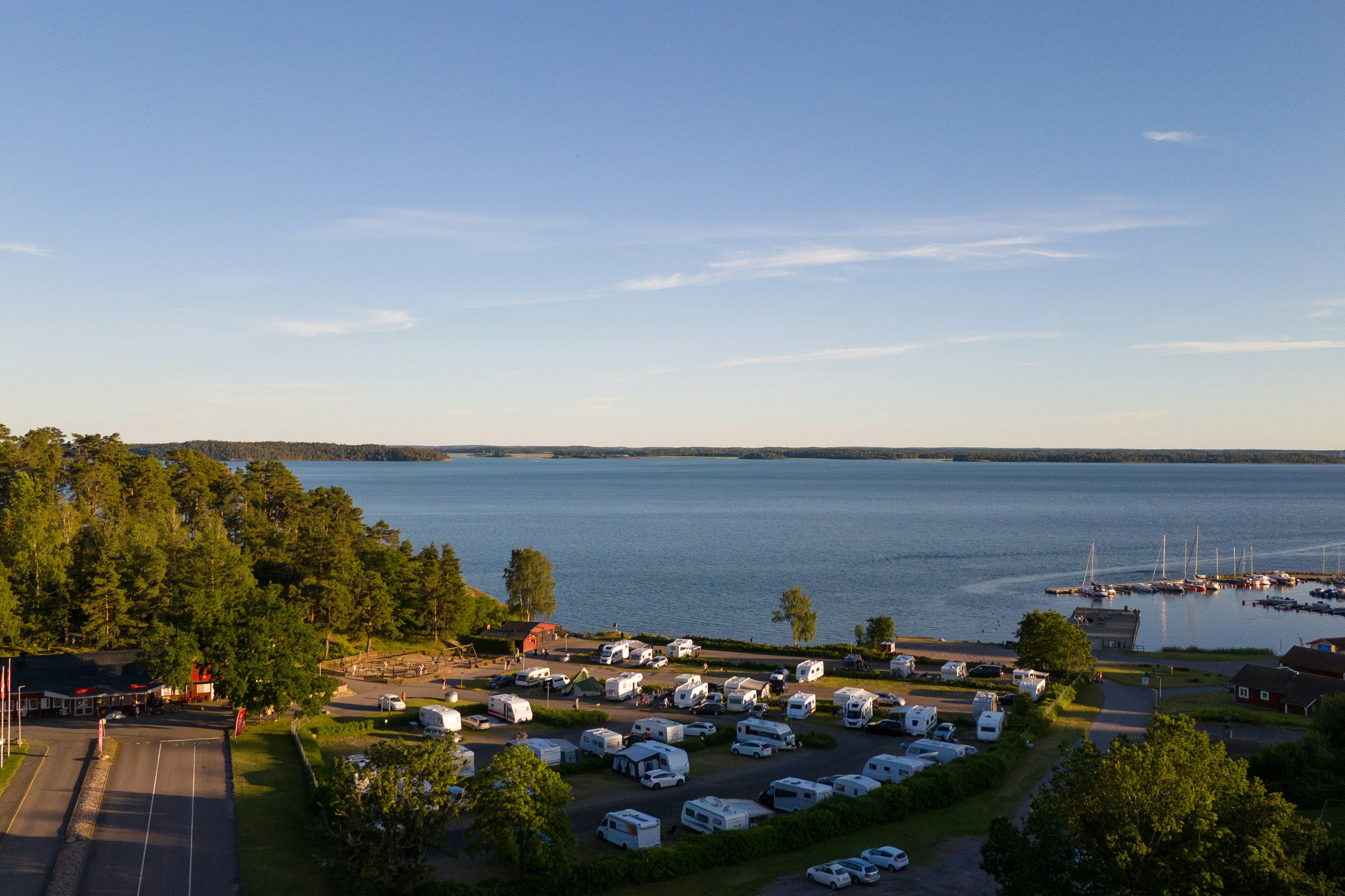 Campingplatz am See mit Wohnwagen und Zelten, Blick auf das Wasser und die Uferlinie, Bäume und Wege im Vordergrund.