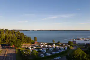 Campingplatz am See mit Wohnwagen und Zelten, Blick auf das Wasser und die Uferlinie, Bäume und Wege im Vordergrund.
