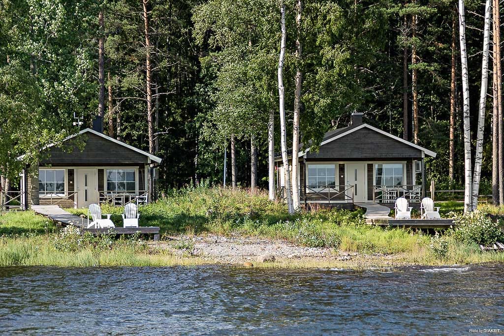 Zwei Campinghäuser am See im Wald, mit Terrasse, Gartenmöbeln und Birkenstämmen im Hintergrund.