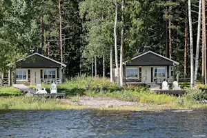 Zwei Campinghäuser am See im Wald, mit Terrasse, Gartenmöbeln und Birkenstämmen im Hintergrund.