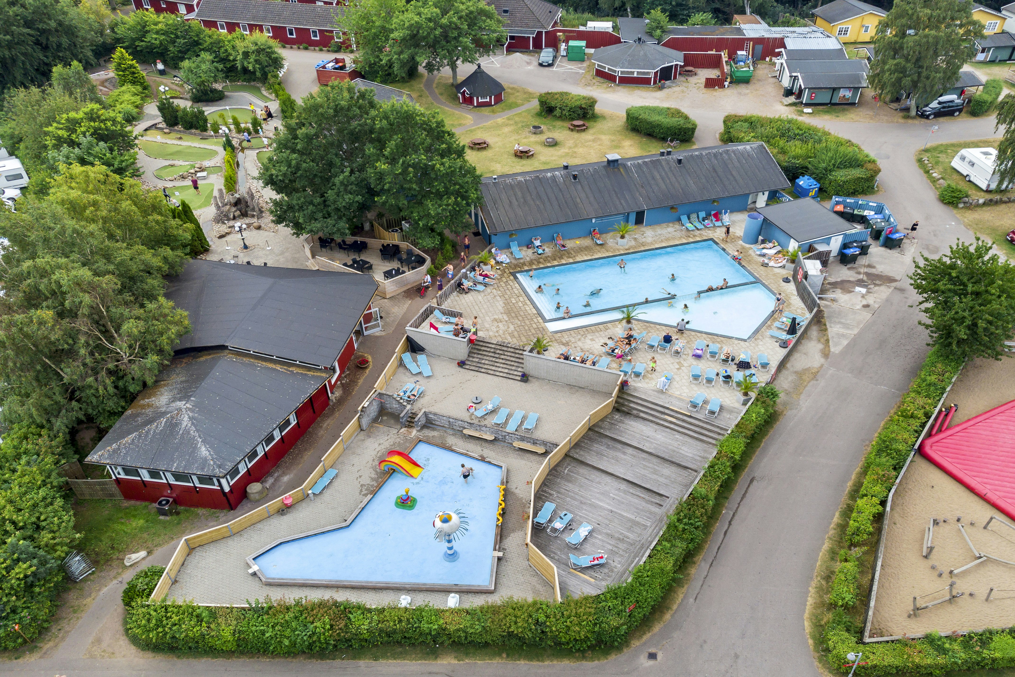 Luftaufnahme eines Campingplatzes mit großem Pool, Liegen, Spielbereichen und mehreren Wohnwagen sowie Gebäuden und Wegen.