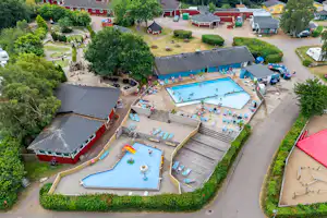 Luftaufnahme eines Campingplatzes mit großem Pool, Liegen, Spielbereichen und mehreren Wohnwagen sowie Gebäuden und Wegen.