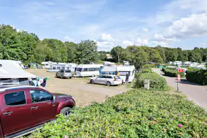 Campingplatz mit Stellplätzen, Wohnmobilen und Autos, Blick über einen Kiesweg und Hecken mit Blumen zum Hintergrund.
