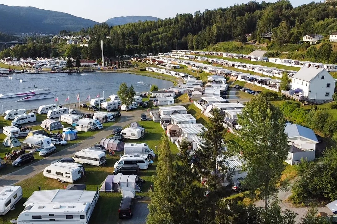 Campingplatz am See mit Wohnwagen und Stellplätzen, Rasenflächen, Booten im Wasser und Häusern am Ufer, umgeben von Wäldern.