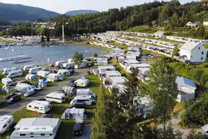 Campingplatz am See mit Wohnwagen und Stellplätzen, Rasenflächen, Booten im Wasser und Häusern am Ufer, umgeben von Wäldern.