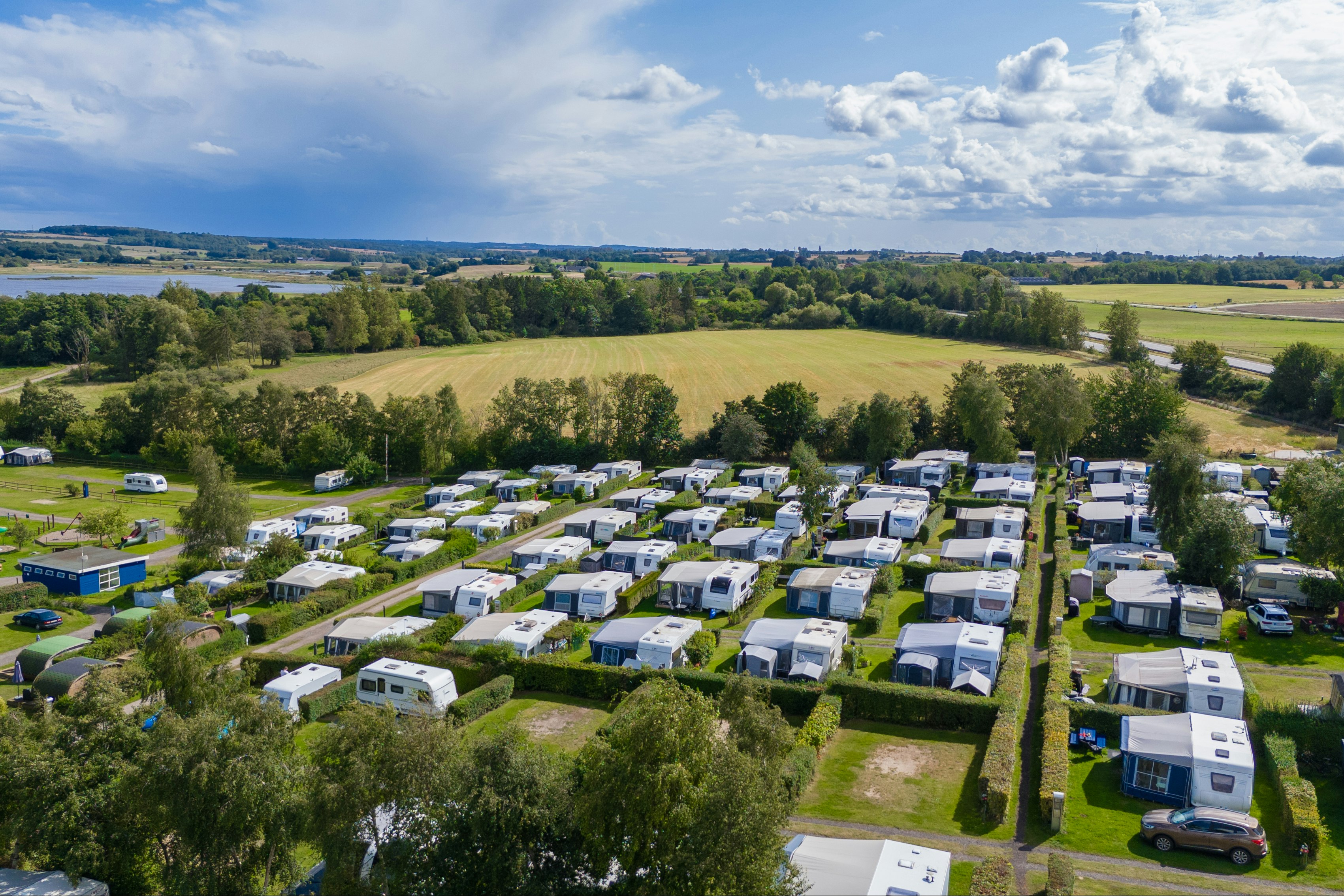 Luftaufnahme eines Campingplatzes mit vielen Wohnwagen und Stellplätzen, Wegen, Rasenflächen und Bäumen in grüner Landschaft.