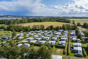 Luftaufnahme eines Campingplatzes mit vielen Wohnwagen und Stellplätzen, Wegen, Rasenflächen und Bäumen in grüner Landschaft.