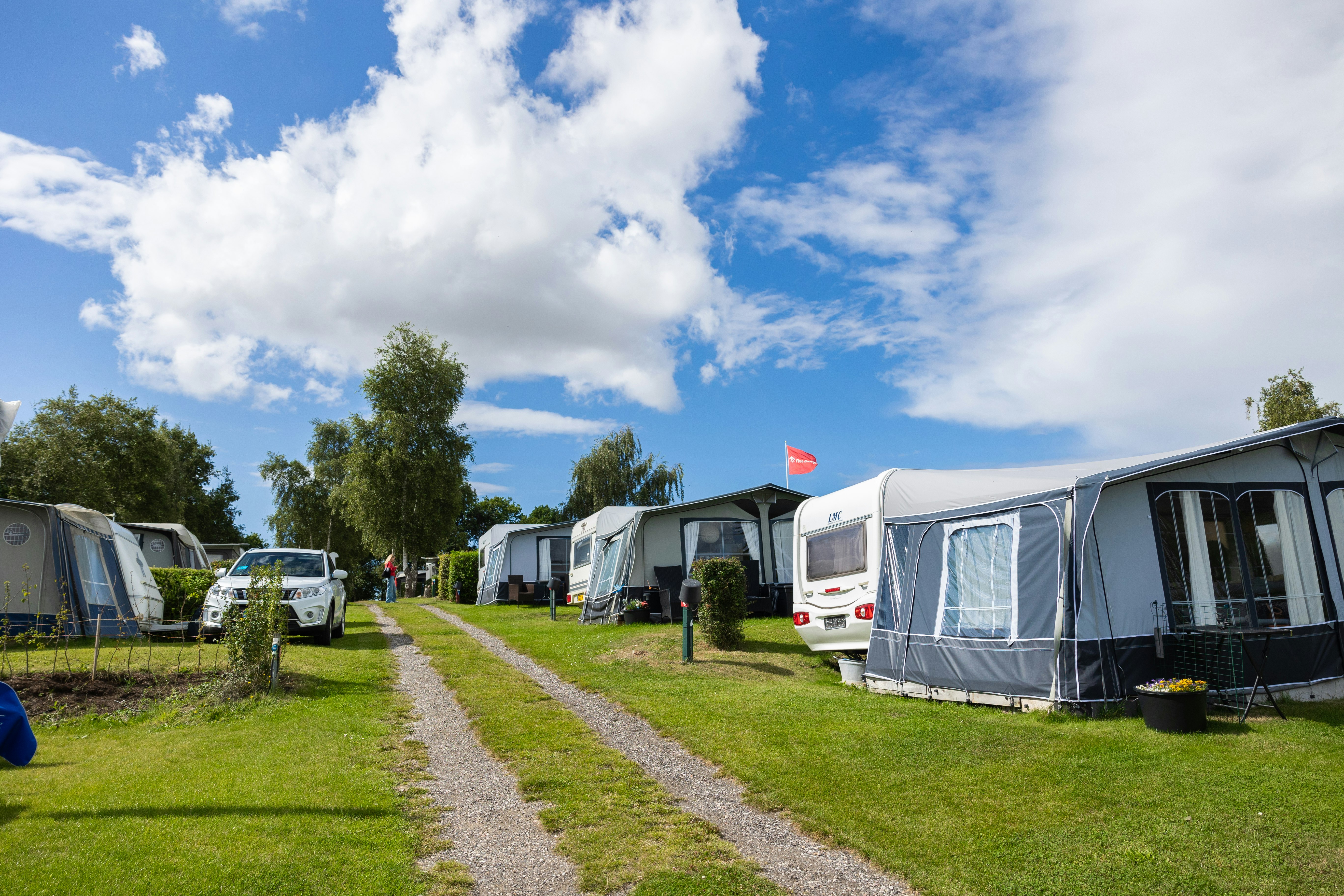 Reihen von Wohnwagen und Zelten auf einem Campingplatz, mit Kiesweg zwischen den Stellplätzen und Bäumen entlang der Wiese.