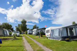 Reihen von Wohnwagen und Zelten auf einem Campingplatz, mit Kiesweg zwischen den Stellplätzen und Bäumen entlang der Wiese.