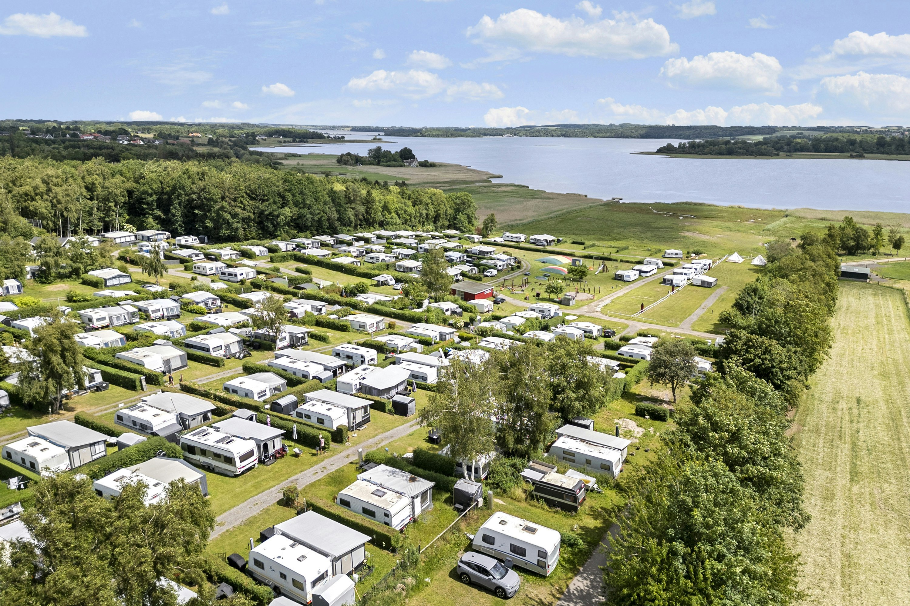 Luftaufnahme eines Campingplatzes mit Wohnmobilen und Stellplätzen, grünen Wegen und einem nahegelegenen See mit Uferlinie.