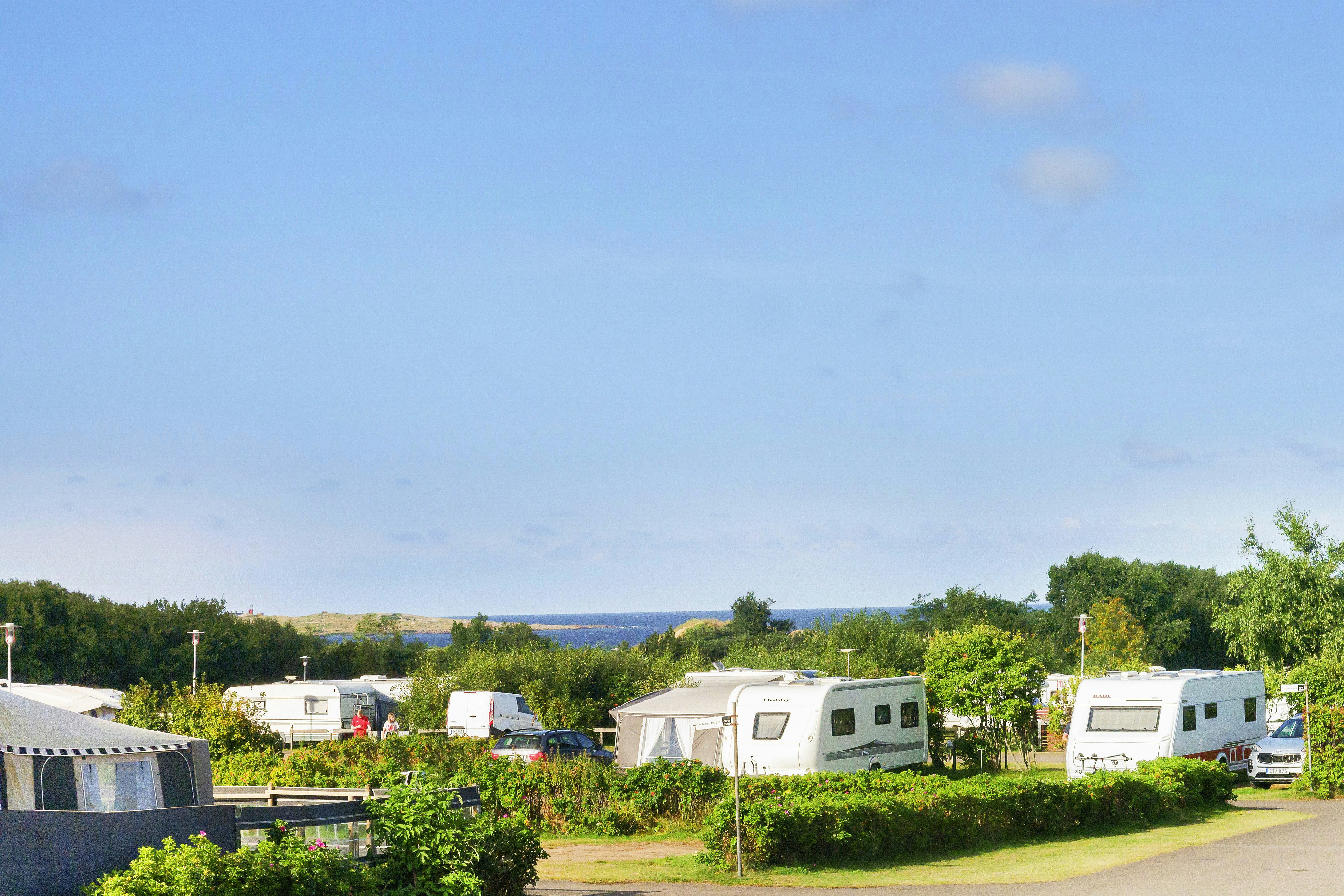 Wohnwagen und Campingfahrzeuge auf einem Stellplatz, Hecken und Bäume im Vordergrund, Straße und Zäune im Hintergrund.