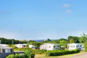 Wohnwagen und Campingfahrzeuge auf einem Stellplatz, Hecken und Bäume im Vordergrund, Straße und Zäune im Hintergrund.