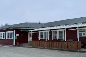 Campinghaus mit Veranda, großen Fenstern und Holzschiebetafel vor dem Eingang, rotes Gebäude mit Metall-Dach.