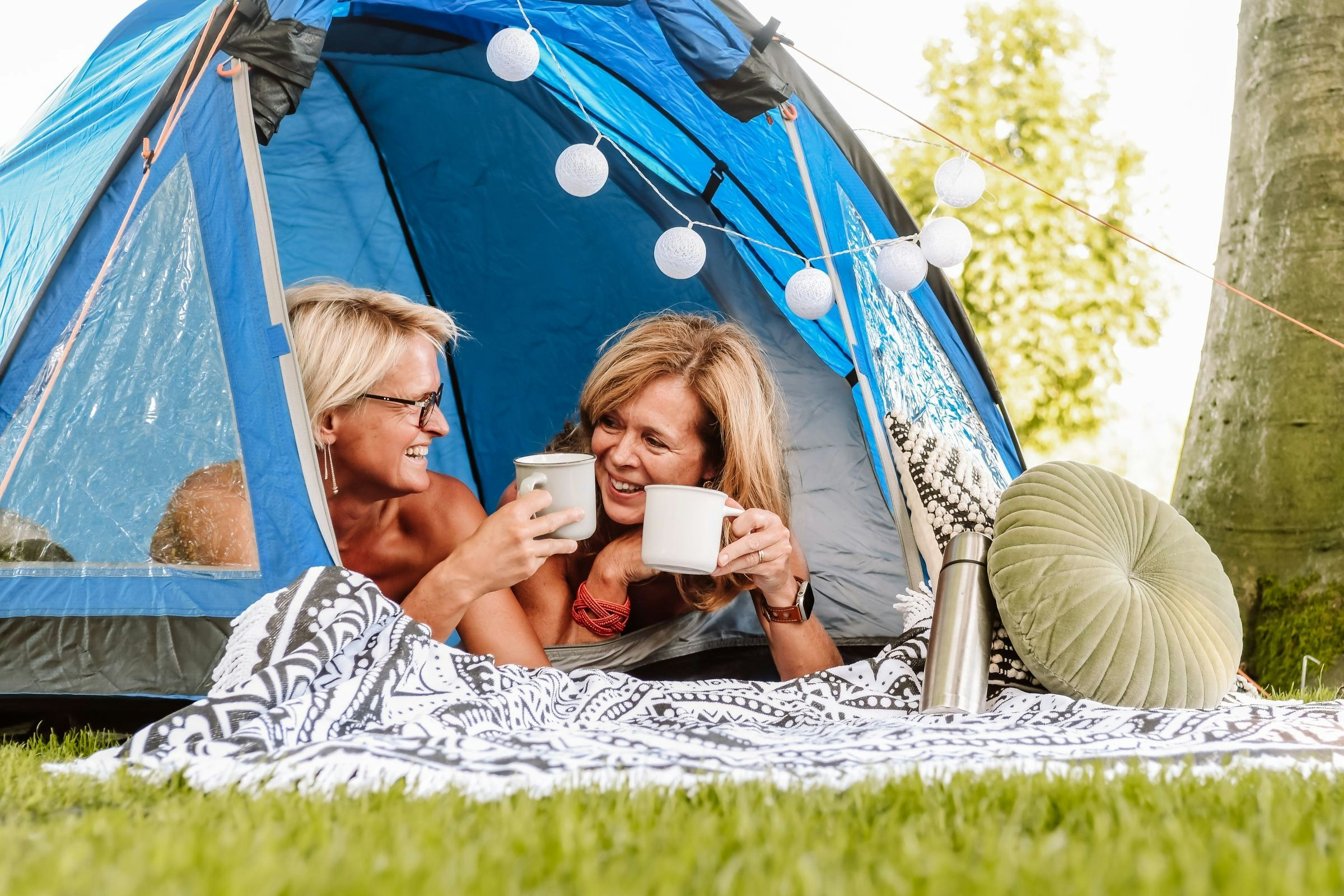 Blaues Campingzelt mit Lichterkette, zwei Personen sitzen davor und halten Kaffeetassen in der Hand.