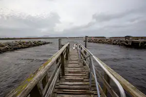 Holzsteg mit Geländern führt ins Wasser, im Hintergrund Hafenmauer und Wellen bei bewölktem Himmel.