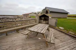 Holzterrasse mit Grillstelle und Holztisch, daneben ein Holzgeländer; im Hintergrund Gebäude und Wiese.
