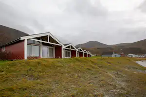 Reihen von Camping-Bungalows mit Terrassen und großen Fenstern, Rasenfläche davor und Hügel im Hintergrund.