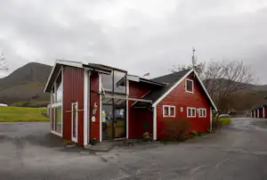 Rotes Gebäude auf einem Campingplatz mit Glasfront, Eingangstür und angrenzendem Parkplatz, im Hintergrund Wiesen und Berge.
