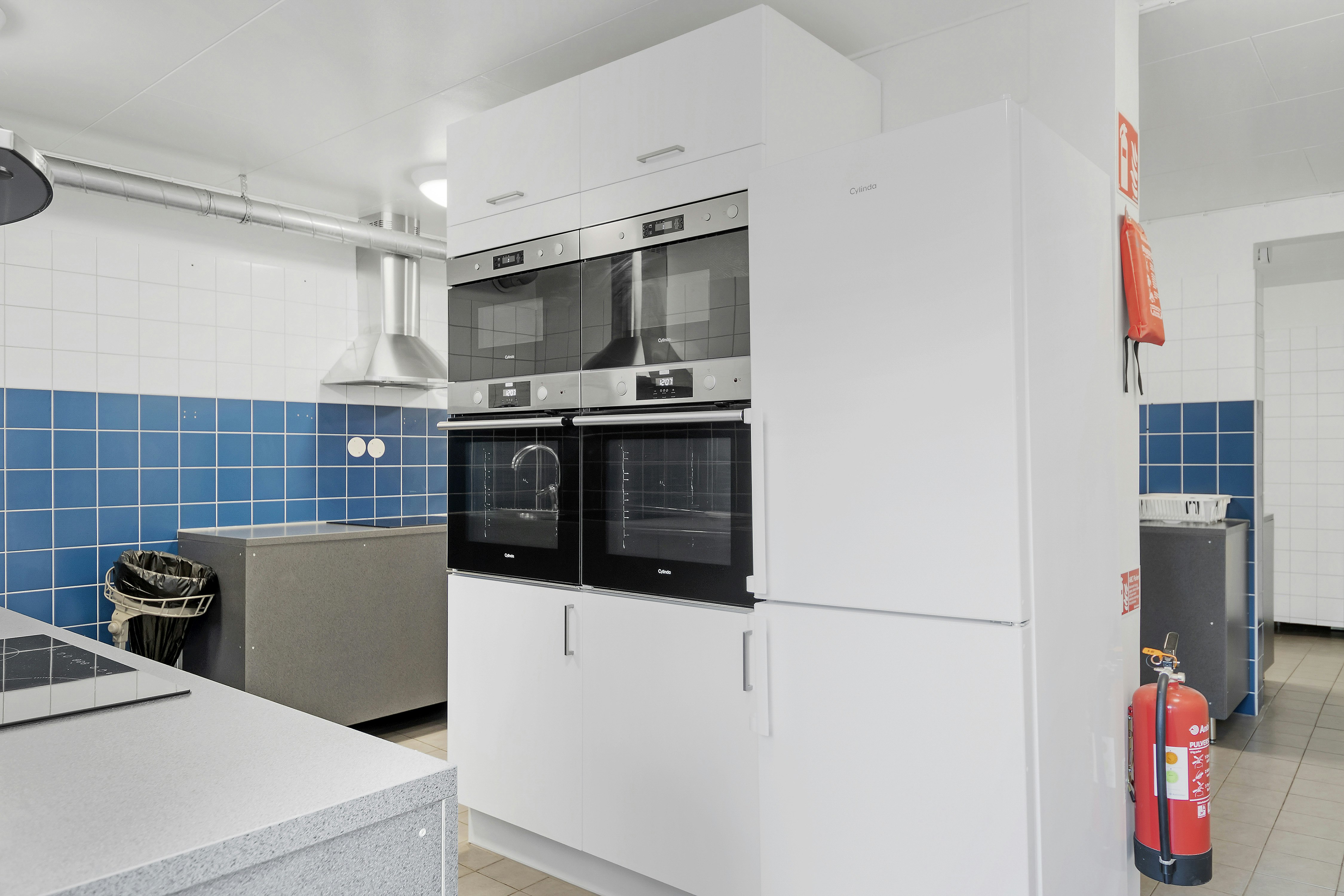 Weiße Küchenzeile mit Backofen und Mikrowelle, Fliesenwand mit blauem Muster, Feuerlöscher und Schrank im Hintergrund.