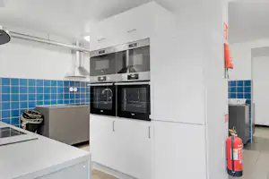 Weiße Küchenzeile mit Backofen und Mikrowelle, Fliesenwand mit blauem Muster, Feuerlöscher und Schrank im Hintergrund.