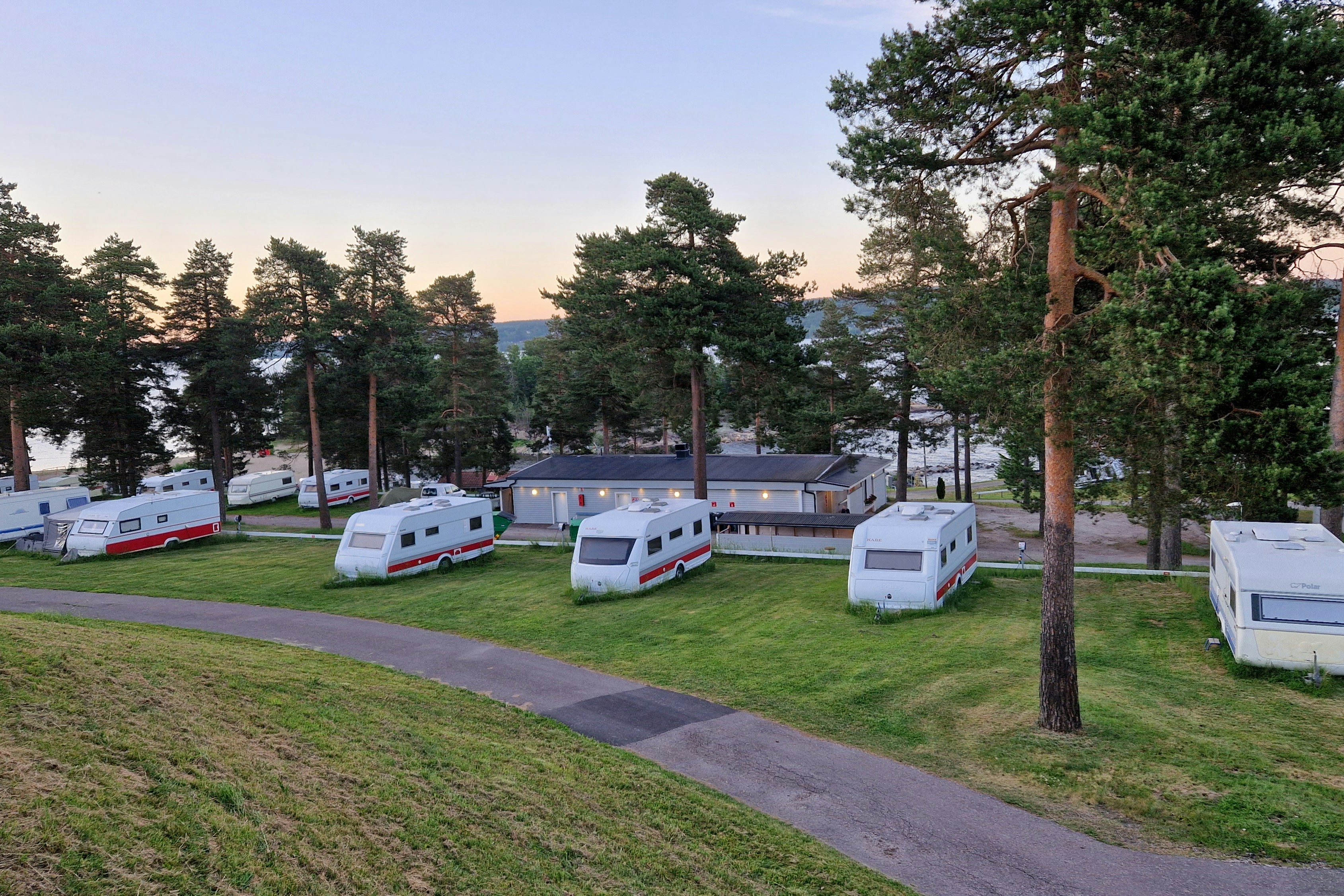 Reihe weißer Wohnwagen auf einem Campingplatz, umgeben von Rasen, Bäumen und einem breiten Weg.