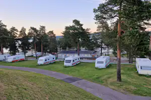 Reihe weißer Wohnwagen auf einem Campingplatz, umgeben von Rasen, Bäumen und einem breiten Weg.