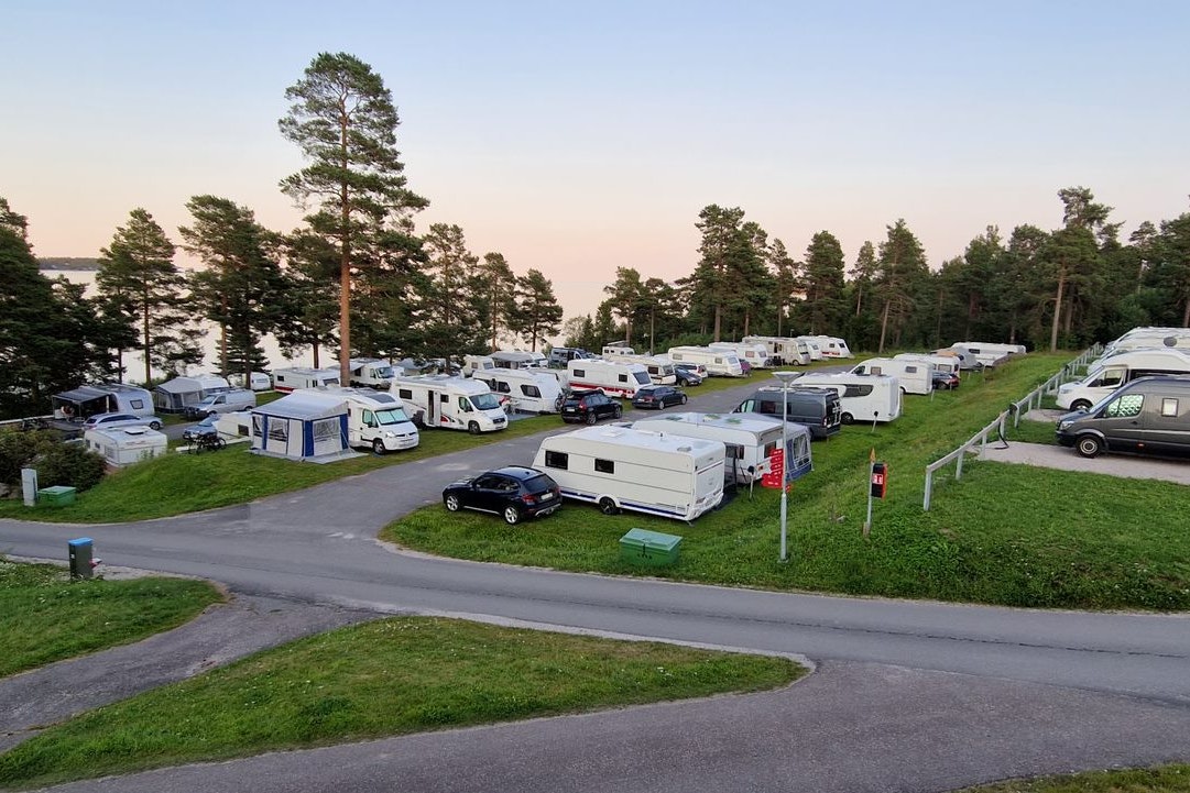 Campingplatz mit Wohnmobilen und Wohnwagen auf Stellplätzen, Rasenflächen und Bäumen entlang der Straße.