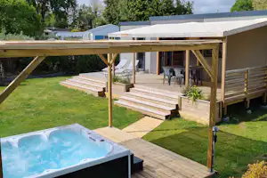 Holzterrasse mit Whirlpool im Vordergrund, Stufen und Sitzbereich unter einem überdachten Anbau, umgeben von Rasen und Bäumen.