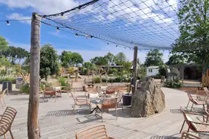 Holzterrasse mit Sitzgruppe und Sonnenschirmen, darüber ein Netzsegel, im Hintergrund Bungalows und Spielplatz im Campingbereich.