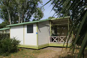 Mobilheim mit Terrasse, Holzstufen und Sitzgelegenheit, umgeben von Bäumen und Sträuchern auf einem Campingplatz.