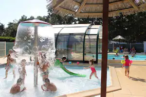 Wasserspiel im Schwimmbecken: Kinder spritzen an einer Fontäne, im Hintergrund ein Poolbereich mit Sonnenschirm und Liegen.
