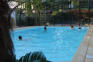 Blauer Campingpool mit Kindern im Wasser, Glasfront und Palmen im Hintergrund, Zaun und Spielbereich sichtbar.