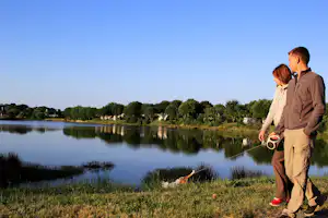 Mann mit Angel steht am Seeufer, im Hintergrund Zelte und Wohnwagen zwischen Bäumen am Campingplatz.