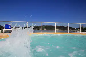 Spritzendes Wasser im Pool vor einer überdachten Glasfront, im Hintergrund Bäume und Gebäude am Ufer.
