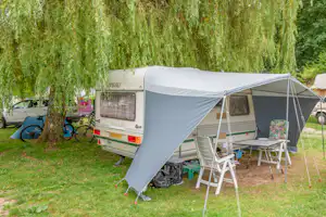 Wohnwagen mit Vorzelt auf einem Campingplatz, daneben Tisch und Stühle, im Hintergrund Bäume und ein Auto.