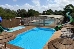 Camping-Pool mit Holzdeck, Glasüberdachung und Rutsche, Liegen am Beckenrand und Bäume im Hintergrund.