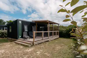 Modernes Campinghaus mit Veranda, zwei Stühlen und Holzpodest im Garten, Hecken und Rasenfläche im Vordergrund.