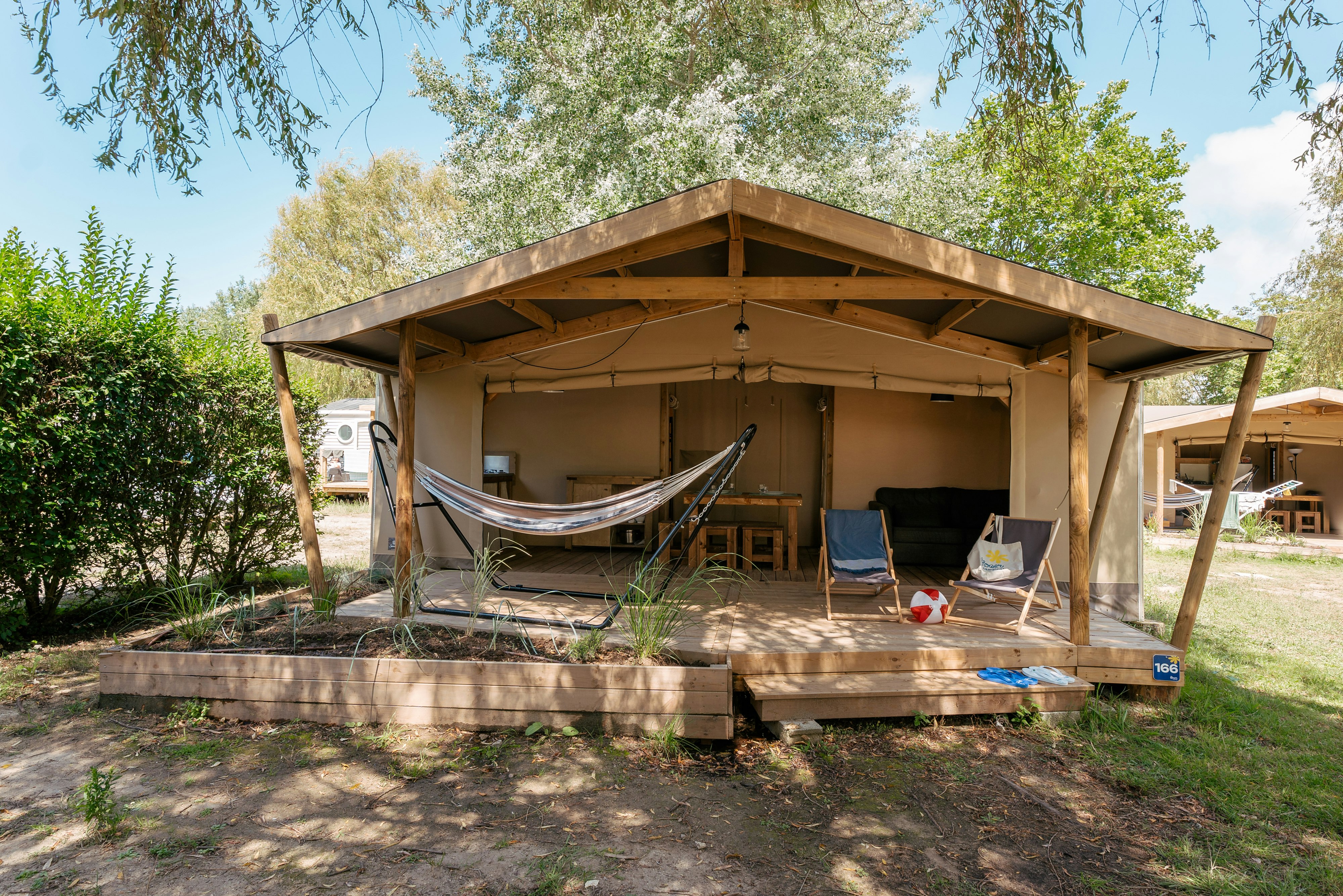 Holzunterstand auf einem Campingplatz mit Hängematte, Sitzgelegenheiten und Palettenboden, umgeben von Bäumen und Rasen.