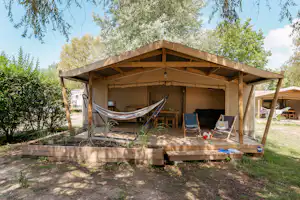 Holzunterstand auf einem Campingplatz mit Hängematte, Sitzgelegenheiten und Palettenboden, umgeben von Bäumen und Rasen.