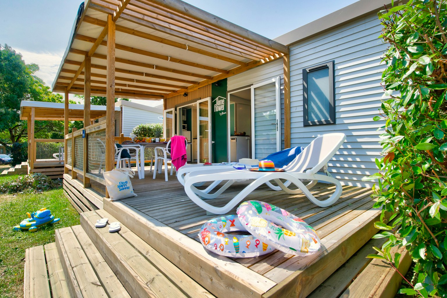 Holzterrasse am Bungalow mit zwei Liegen, Sitzbank, Tisch und Blick auf den Eingang, umgeben von Büschen und Rasen.