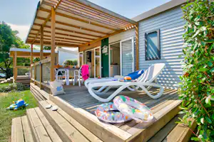 Holzterrasse am Bungalow mit zwei Liegen, Sitzbank, Tisch und Blick auf den Eingang, umgeben von Büschen und Rasen.