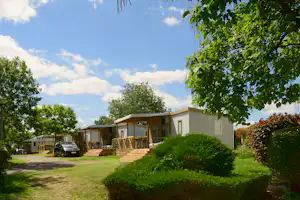 Wohnwagen und Mobilheime mit Holzterrasse, Rasenweg und Büschen im Campingplatz, umgeben von Bäumen und Himmel.