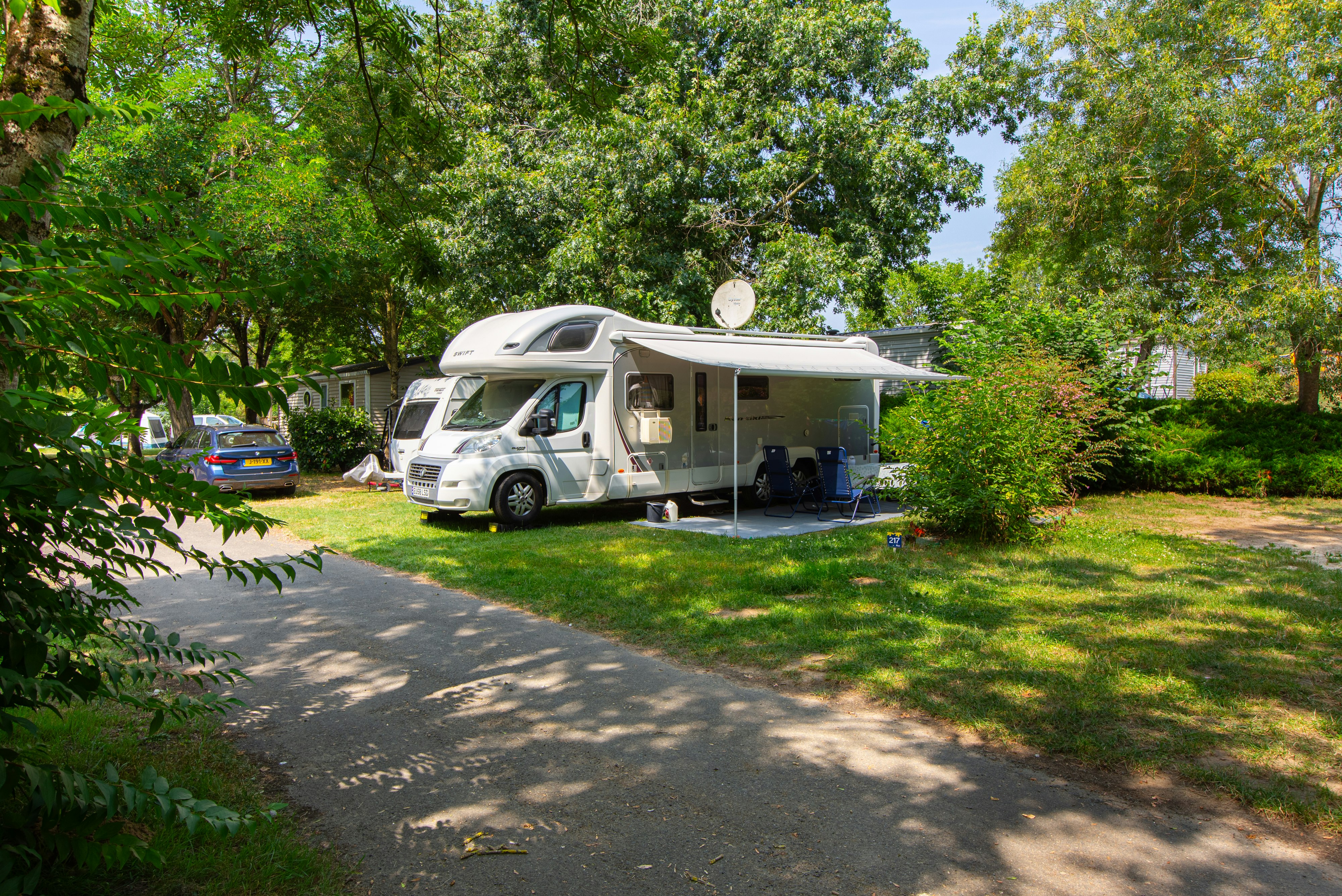 Wohnmobil auf einem Stellplatz im Grünen, mit Vorzelt und Tisch, umgeben von Bäumen und einem gepflasterten Weg.
