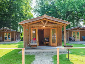 Holz-Campinghaus mit Veranda, Sitzbank und Tisch, im Grünen zwischen weiteren Holzhütten und Rasenfläche.