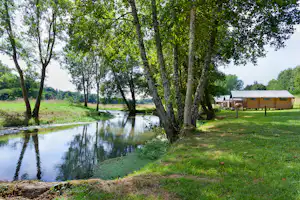 Wohnwagen am Flussufer im Grünen, mit Holzterrasse und Sitzbereich, umgeben von hohen Bäumen und Rasen.