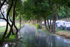 Ruhiger Fluss am Campingplatz, gesäumt von Bäumen; rechts stehen Wohnwagen und ein Weg führt am Ufer entlang.