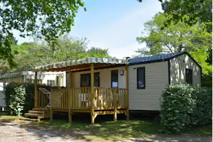 Campinghaus mit Holzveranda, Markise und großen Fenstern, umgeben von Bäumen und Hecken im grünen Außenbereich.