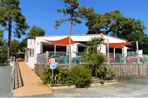 Camping-Unterkunft mit Terrasse und orange-weißem Zeltdach, Holzgeländer, Pflanzen im Vordergrund und Bäume im Hintergrund.