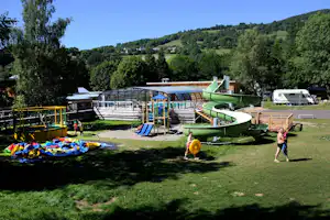 Spielplatz auf dem Campingplatz mit Rutsche, Klettergerüst und Trampolin, im Hintergrund Wohnwagen und Bäume vor grünen Hügeln.