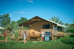Großes Campingzelt auf Holzpodest mit Grill, Sitzbank und Gartenmöbeln, umgeben von Palmen und grünem Rasen.